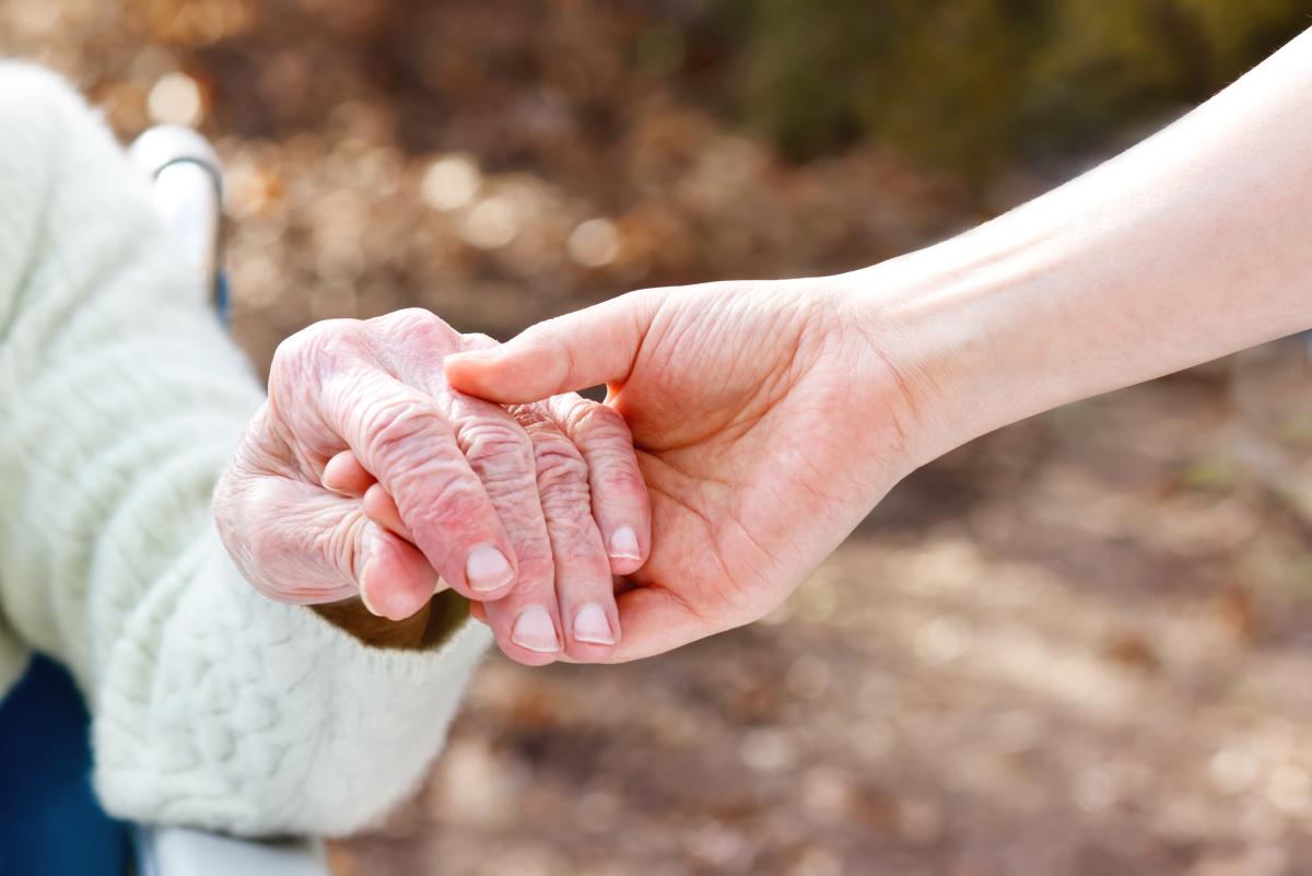 Die Hand einer jungen Person hält unterstützend die Hand einer älteren Person, die im Rollstuhl sitzt.