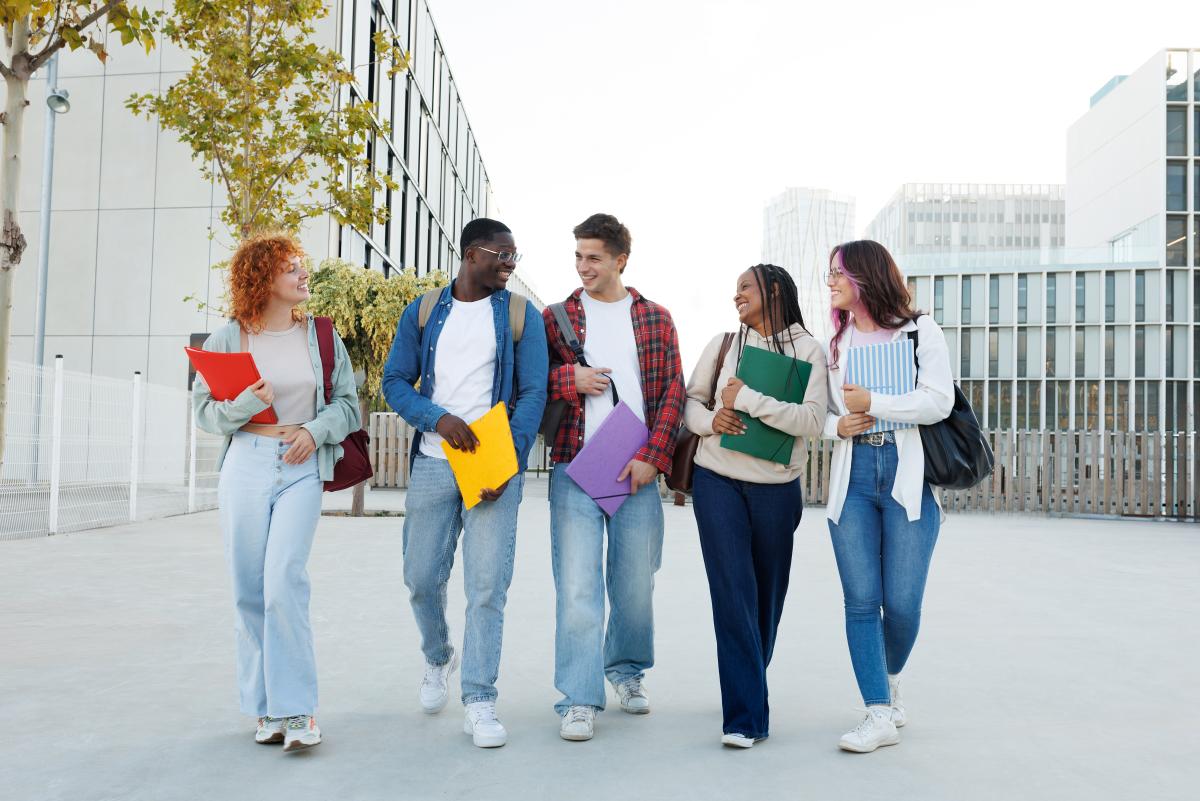 Fünf Studierende mit Heftern spazieren lachen und sich unterhaltend über den Campus.